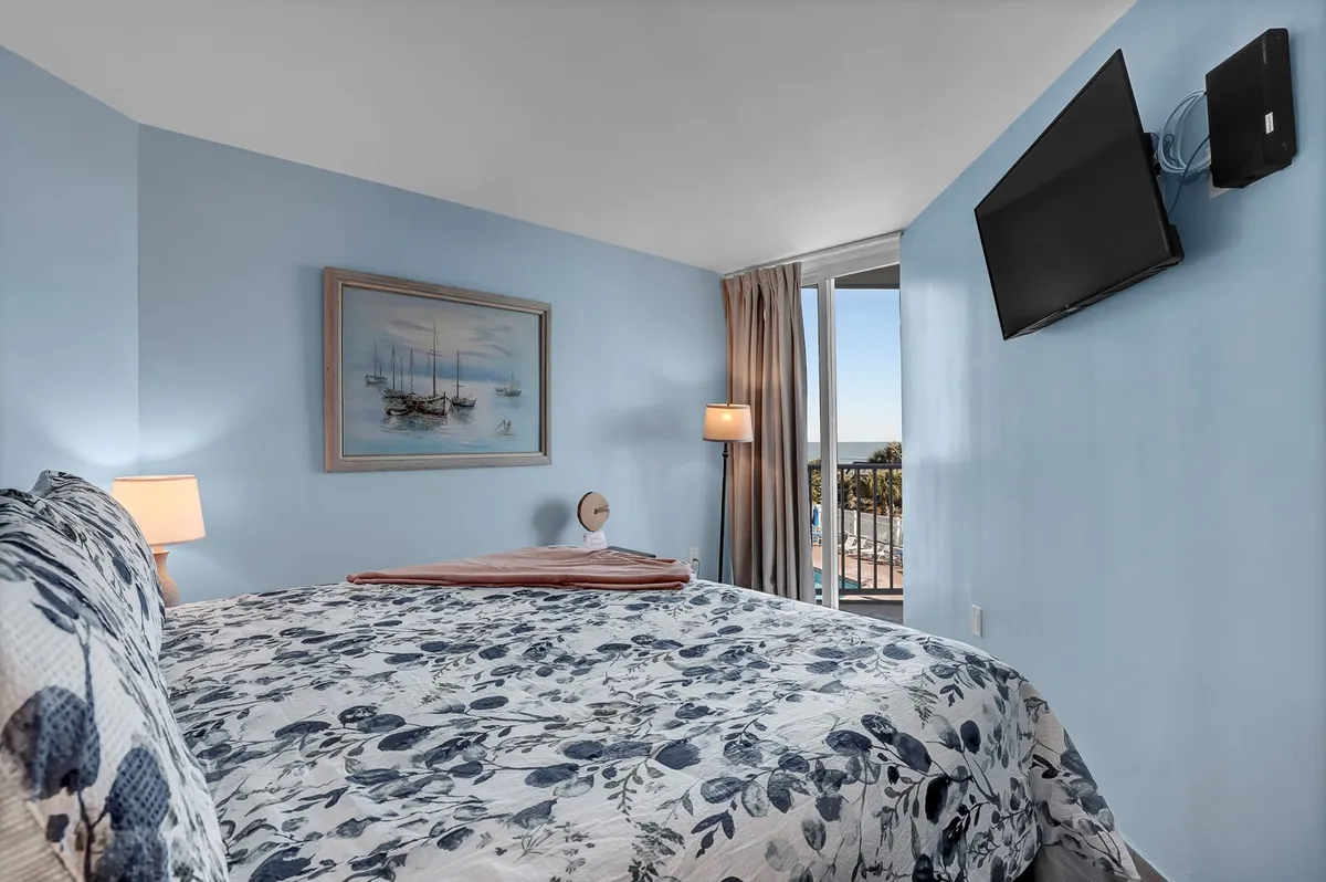 A sun-filled bedroom with a bed featuring floral bedding, a framed painting of boats above the headboard, floor-to-ceiling windows letting in natural light, and a warm bedside lamp. The decor is calm and serene, with a distinct coastal influence.