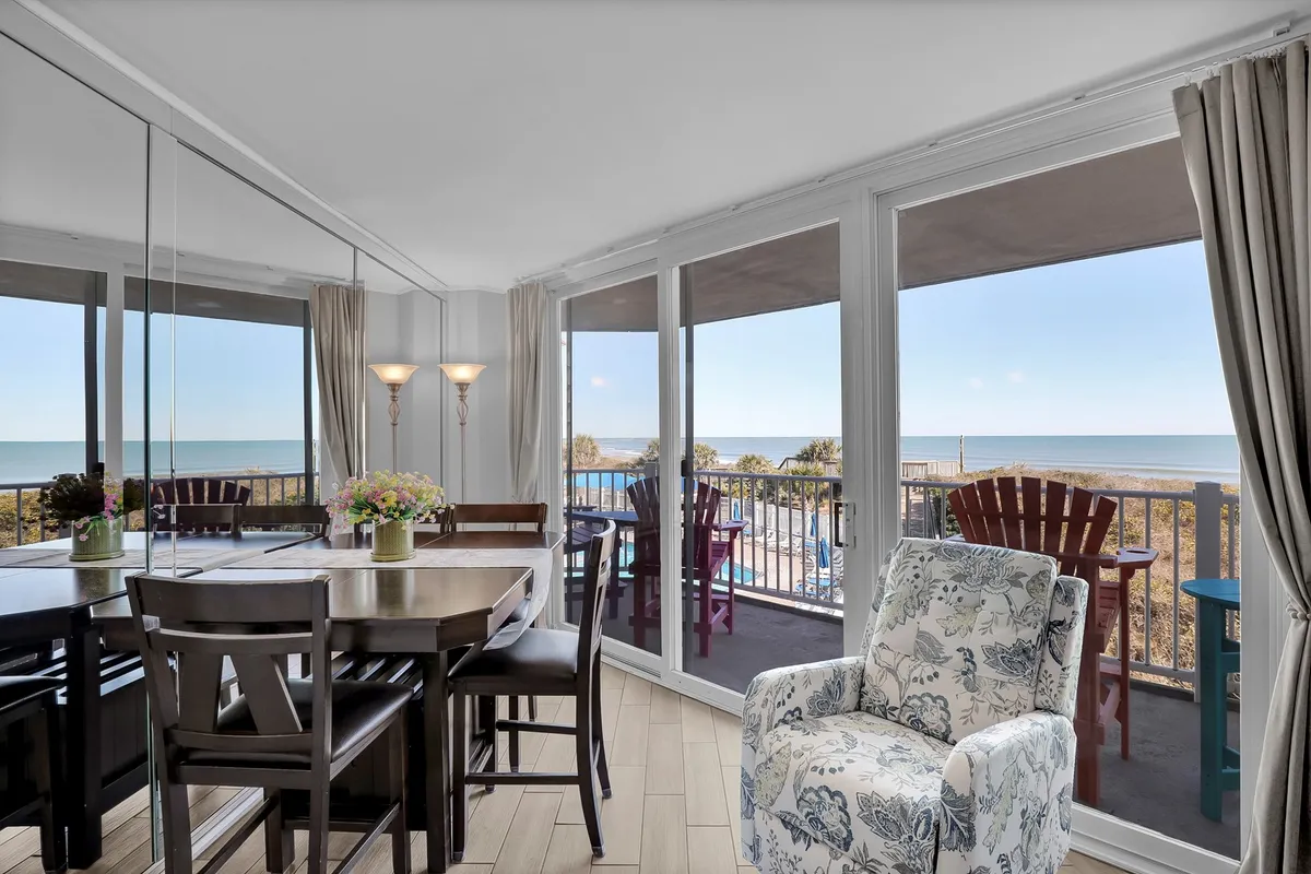 The dining table sits beside expansive windows with an uninterrupted view of the ocean, accompanied by a comfortable chair that invites reading or conversation. Soft, natural light fills the space, enhancing its relaxed atmosphere.