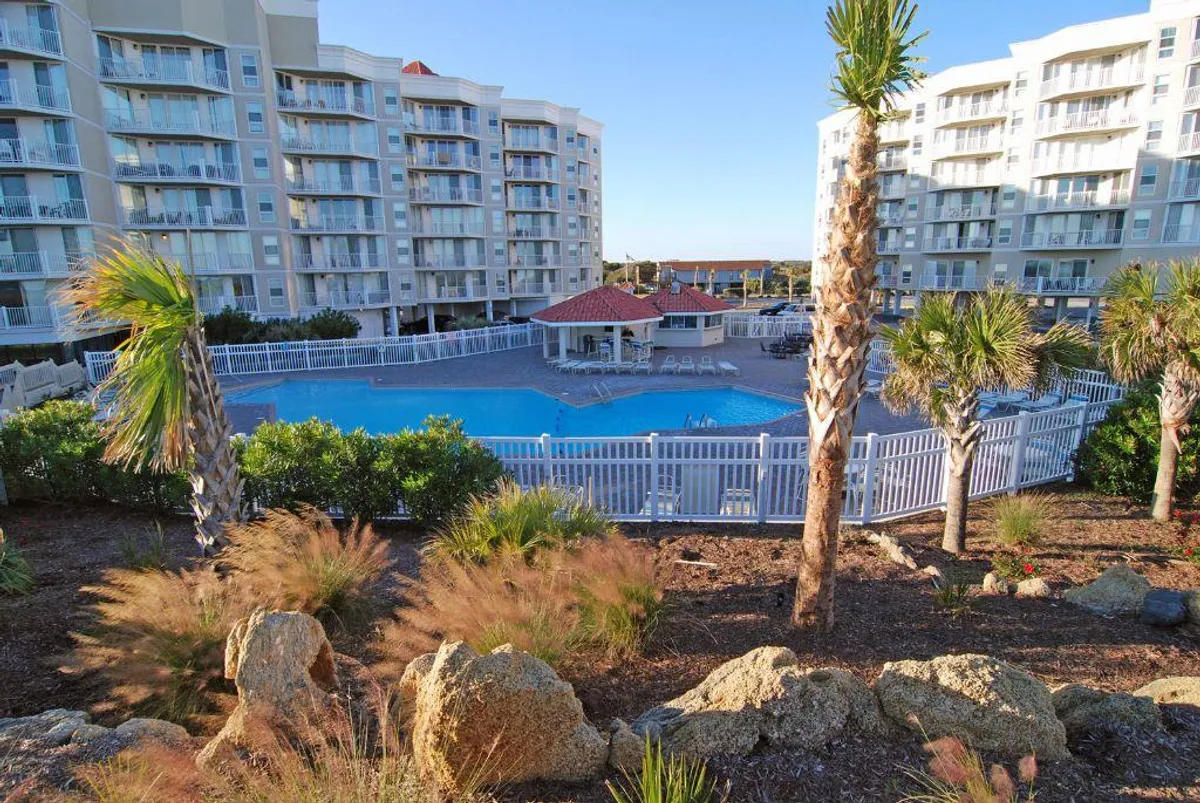 A vibrant outdoor swimming pool surrounded by palm trees and residential buildings, with a poolside gazebo offering shaded seating. The area is well-maintained, inviting for both relaxation and social gatherings, and showcases the kind of leisurely activities available year-round.