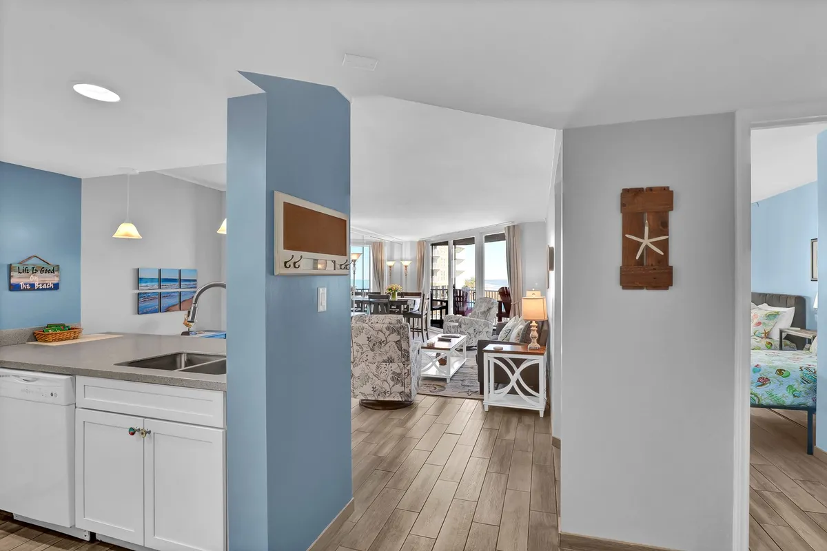 A wide angle reveals the integrated kitchen, dining, and living areas with decorative wall art and a clear view through large windows to the outdoors. The space feels welcoming, bright, and ideal for both meals and relaxation.