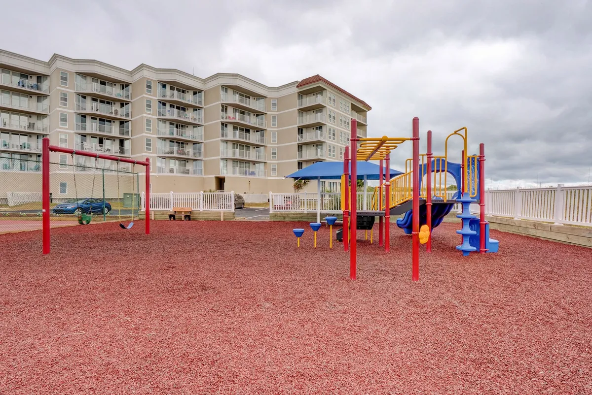 A lively playground with swings and vibrant equipment sits on a cheerful red rubber surface, busy with children and families enjoying the outdoors. An apartment building frames the background, emphasizing the communal, family-friendly aspect of the resort.