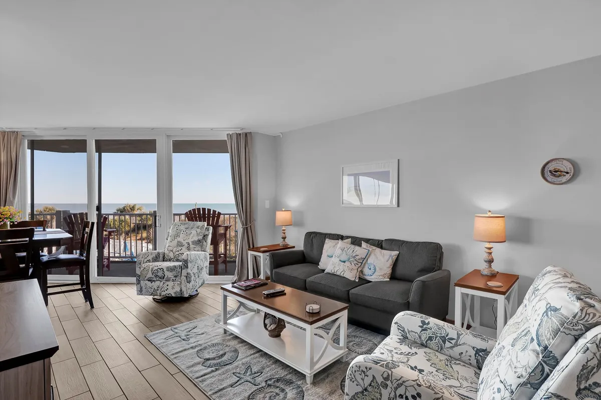 This image showcases a welcoming living and dining area with a comfortable sofa, dining table and chairs, and expansive windows providing a sweeping ocean view. Decorative lamps and a tranquil, beach-inspired color palette enhance the sense of relaxation and togetherness.