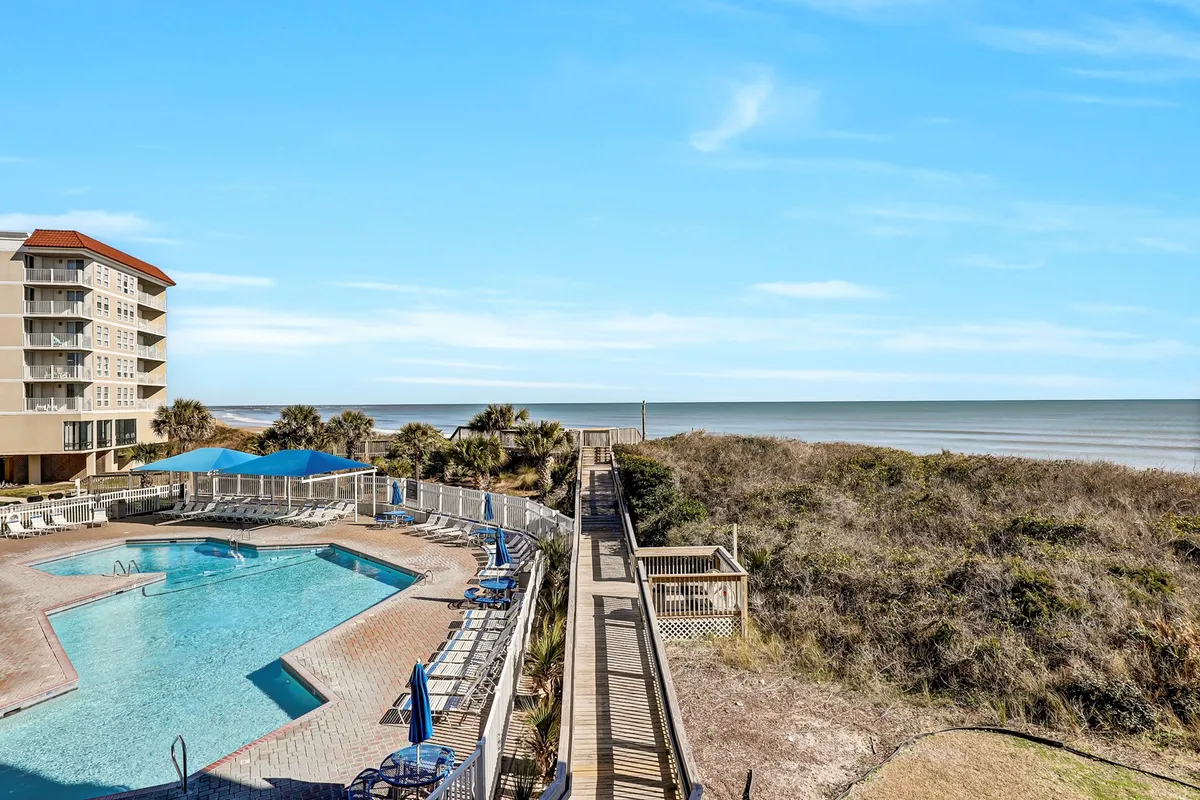 A sparkling swimming pool is set beside a sandy beach and boardwalk, with the condominium building in view. Palm trees and lounge chairs line the pool area, creating a relaxing, resort-like environment ideal for sunbathing or swimming. The ocean is visible just beyond the pool, providing a seamless blend of poolside and beachfront atmospheres.