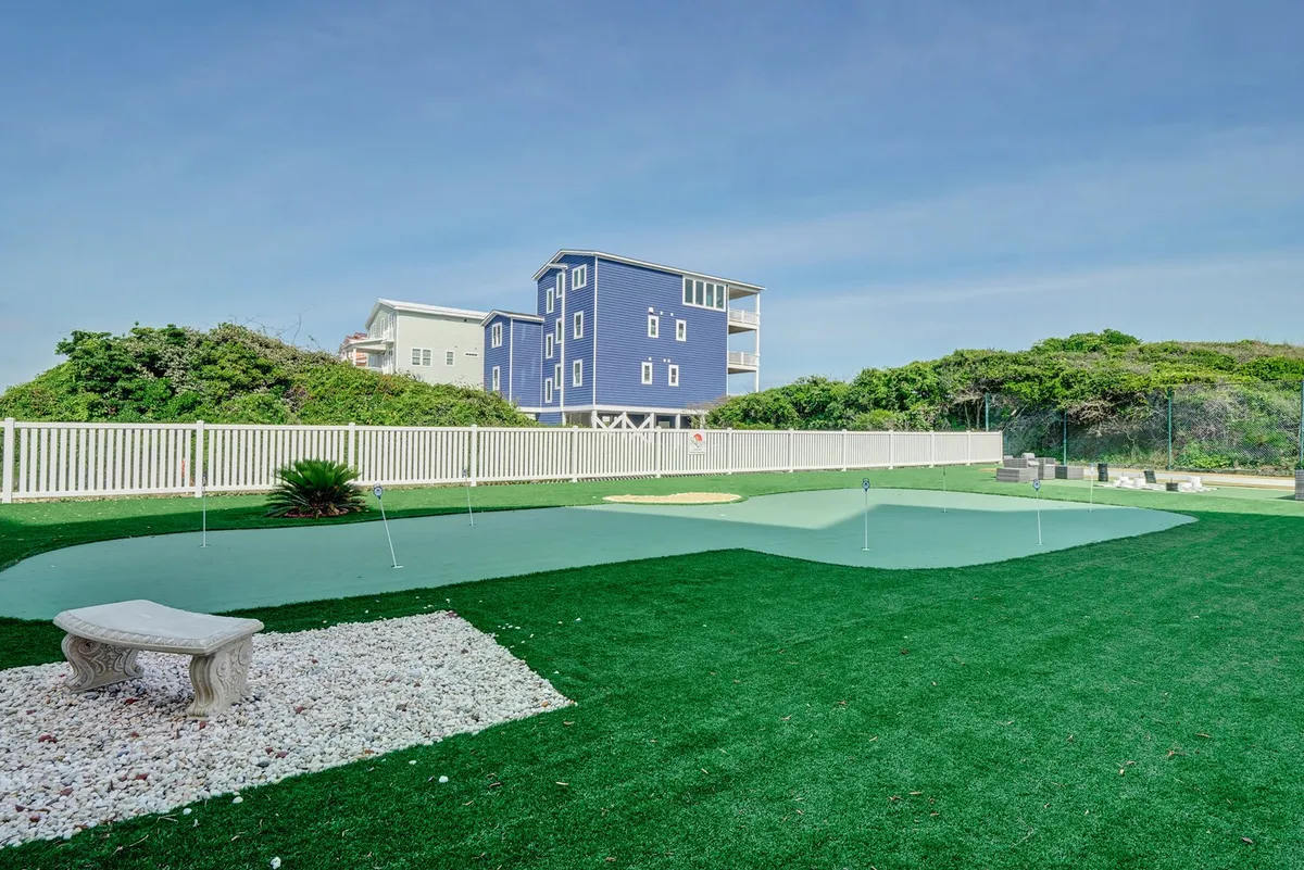 This exterior image features a lush green putting green bordered by white stones, surrounded by landscaping and with a blue house visible in the background. The open, sunlit space invites guests to practice golf or enjoy social outdoor activities.