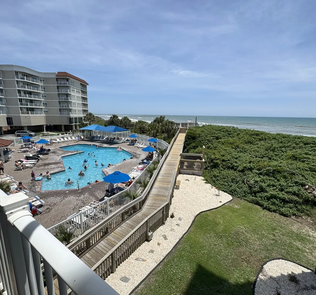 The image showcases the exterior of the St Regis 2209 condo at North Topsail Beach. In the foreground, a sparkling new covered saltwater pool invites guests to swim and relax. Lounge chairs and a deck area are positioned for sunbathing, while a paved pathway leads directly from the pool area to the sandy beach, visible with rolling ocean waves in the background. The sky is painted with hues of sunset, casting a golden glow over the entire scene and creating a tranquil, vacation-ready atmosphere.