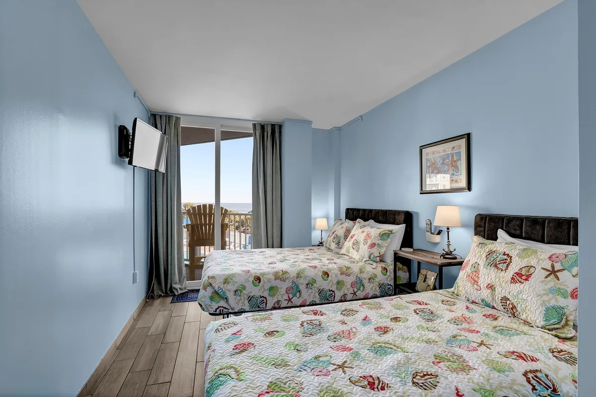 A cheerful bedroom featuring two beds with colorful quilts, a large window showcasing a sea view, and side tables with lamps. The room is bright, playful, and inviting, perfect for children or friends sharing a space.