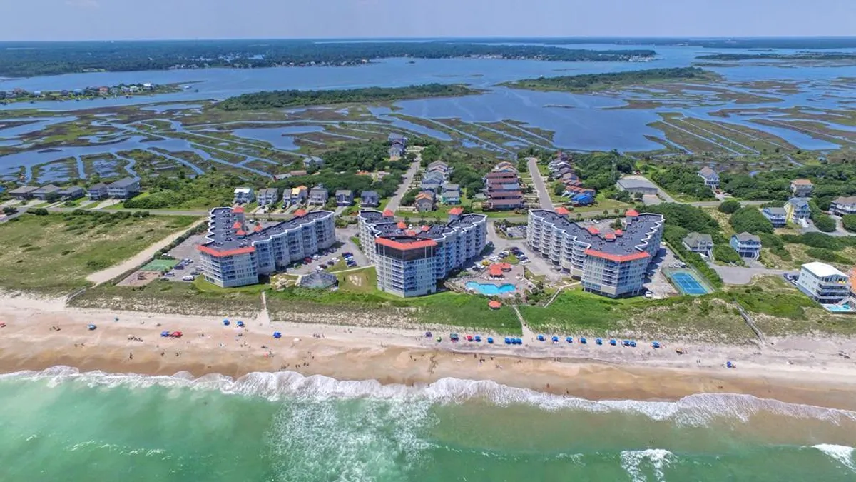 This exterior image features beachfront condominium buildings with a sweeping view of the sandy beach, glistening swimming pool, and ocean waves lapping the shore. Vacationers can be seen enjoying the pool, relaxing on sun loungers, and strolling along the sand, all set against a backdrop of dreamy sunset skies.