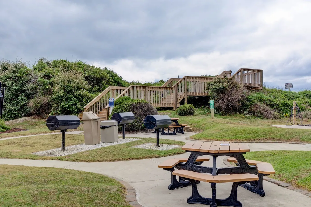 A welcoming barbecue area with picnic tables and barbecue grills set amidst casual surroundings, ideal for family gatherings or friendly get-togethers. A staircase leads upwards, hinting at more amenities nearby and encouraging exploration.