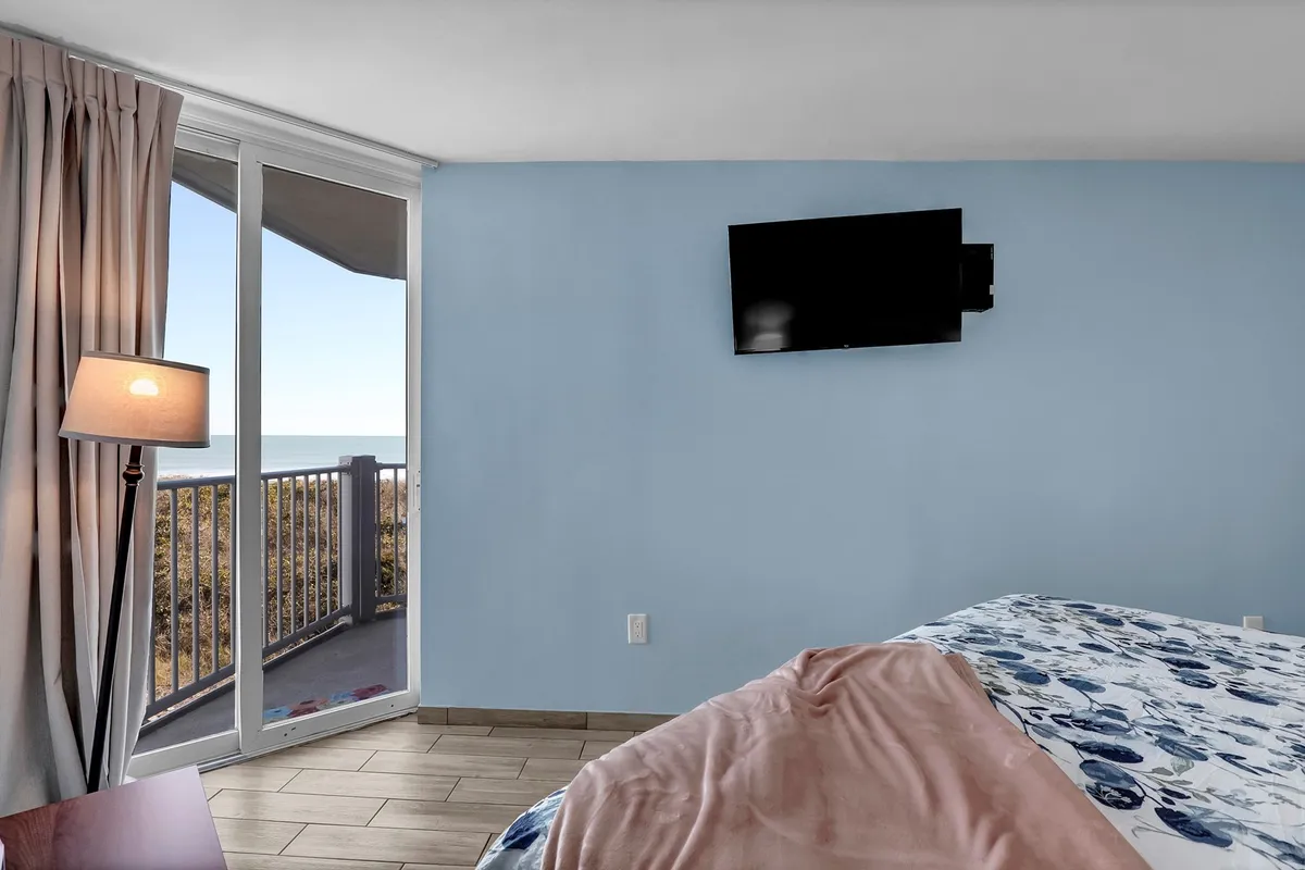 An inviting bedroom featuring a plush bed with floral bedding, a lamp, a TV, and a sliding glass door that opens onto a private balcony overlooking the ocean. The soft natural light and calming decor set a tranquil, intimate mood.