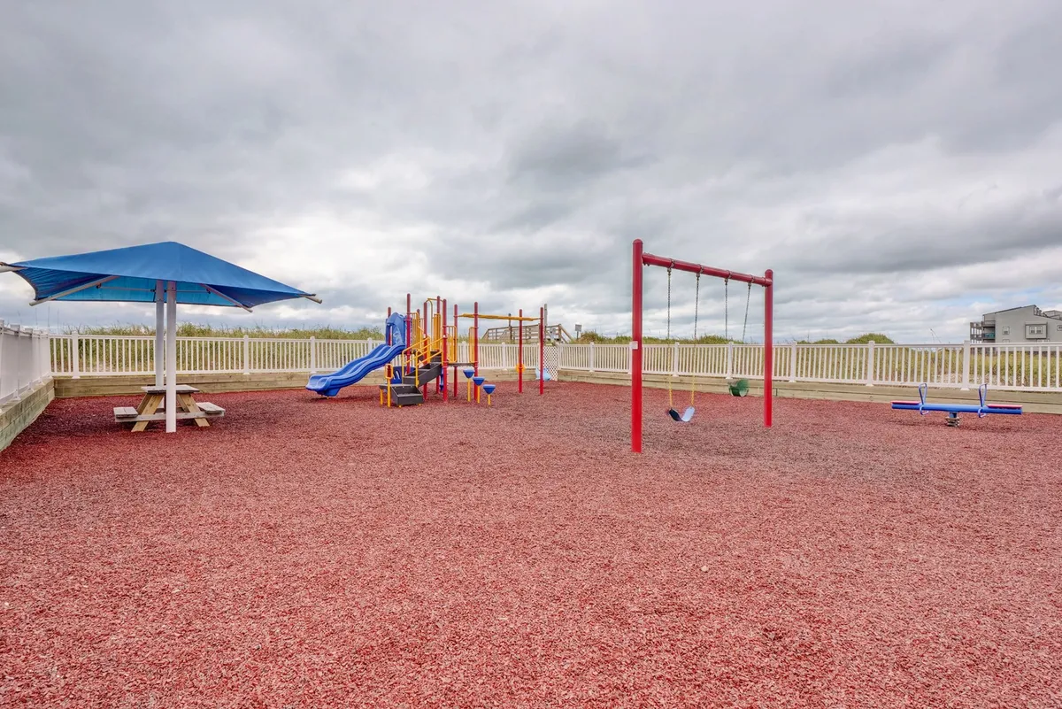 A cheerful playground scene shows vibrant equipment, swings, and a shaded picnic table, inviting families and children to play together. The area is surrounded by sand and a shade structure, creating a safe and joyful environment for kids to explore.